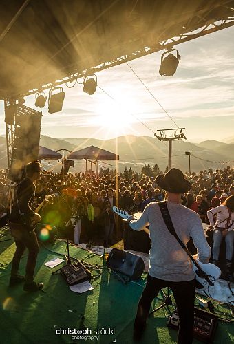Band speelt op buitenpodium in de bergen bij zonsondergang, omringd door groot publiek en bergachtergrond.