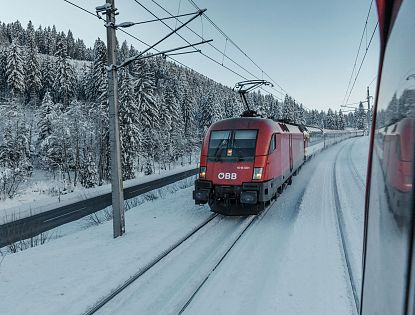 A red ÖBB train travels through a snowy forest landscape with evergreen trees and a road alongside. The area is blanketed in snow creating a wintery scene.