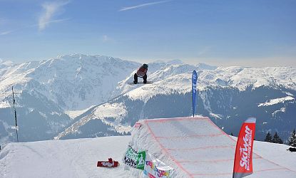 Ein Snowboarder in der Luft führt einen Trick über eine Rampe in einem verschneiten Gebirgspanorama mit blauem Himmel und Bergkette im Hintergrund aus.