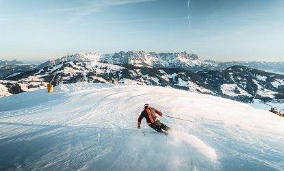 Skiër in feloranje pak descendert van besneeuwde berghelling met majestueus berglandschap op de achtergrond.