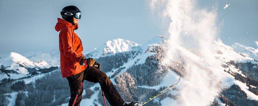 Ein Skifahrer in oranger Jacke und schwarzem Helm steht auf einem schneebedeckten Berg, umgeben von verschneiten Gipfeln und wirbelndem Schnee.