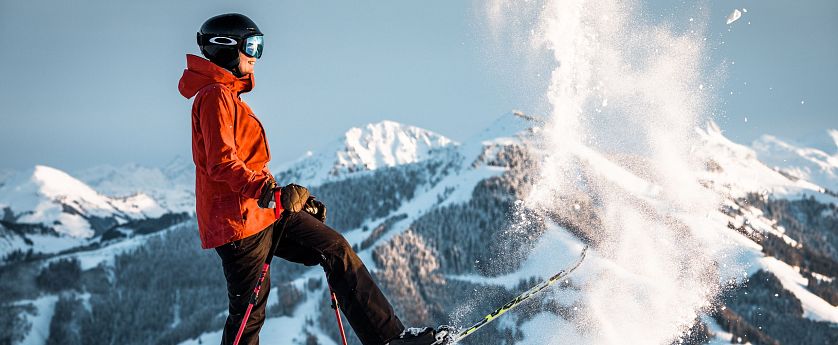 Ein Skifahrer in oranger Jacke und schwarzem Helm steht auf einem schneebedeckten Berg, umgeben von verschneiten Gipfeln und wirbelndem Schnee.