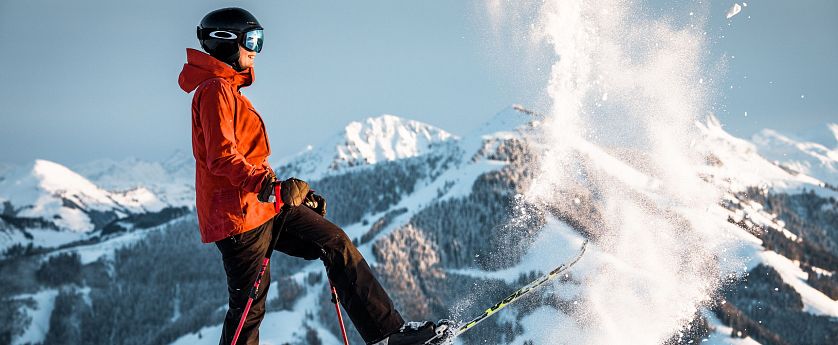 Ein Skifahrer in oranger Jacke und schwarzem Helm steht auf einem schneebedeckten Berg, umgeben von verschneiten Gipfeln und wirbelndem Schnee.