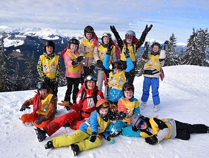 A group of smiling children in colorful ski gear play in the snow, with trees and mountains in the background, enjoying a fun day on the slopes.