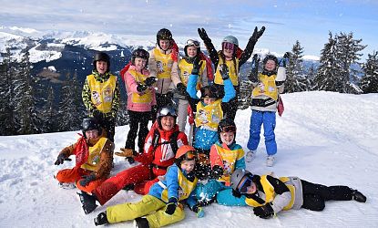 Kinderen in kleurrijke skikleding spelen lachend in de sneeuw aan de voet van een besneeuwde berg, met skihelmen en zonnebrillen op.