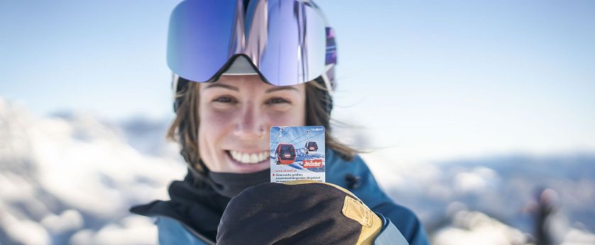 Eine Person in Ski-Ausrüstung hält ein Ticket für eine Bergbahn. Im Hintergrund sind Schnee und ein klarer Himmel zu sehen.
