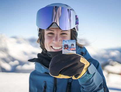 Person mit Skihelm und Schutzbrille hält eine Skipass- oder Werbekarte in der Hand. Im Hintergrund sind Gondeln auf einem verschneiten Berg sichtbar.