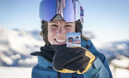 Person mit Skihelm und Schutzbrille hält eine Skipass- oder Werbekarte in der Hand. Im Hintergrund sind Gondeln auf einem verschneiten Berg sichtbar.