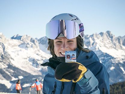 Person in Skikleidung hält ein Ticket für eine Seilbahn. Im Hintergrund verschneite Berge und ein blauer Himmel.
