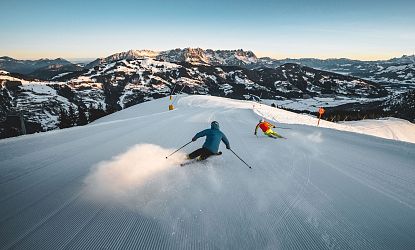 Zwei Skifahrer rasen eine schneebedeckte Piste hinunter, umgeben von majestätischen Bergen. Der Himmel ist klar, und die wechselnden Lichter der Dämmerung schaffen eine zauberhafte Atmosphäre.