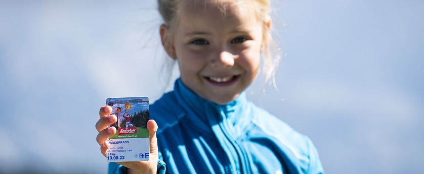 Kind met blauwe jas toont glimlachend een skikaart tegen een wazige blauwe lucht aan.