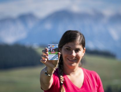 Frau hält eine Karte mit Berglandschaft im Hintergrund. Die Karte hat ein Bild eines Wanderers und ein Datum darauf.