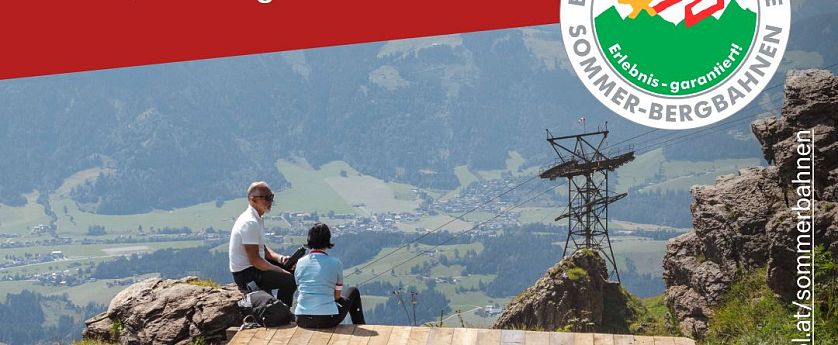 Two people on a wooden platform enjoying a mountain view with a cable car structure and a valley village in the background.