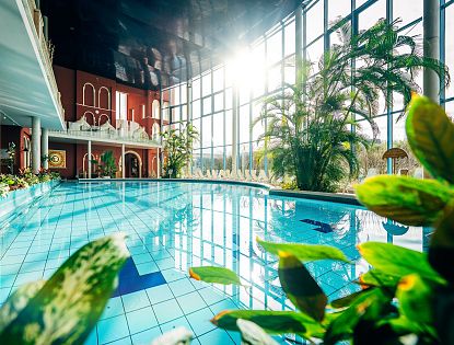 Helles, geräumiges Schwimmbad mit großen Fenstern und üppigen Pflanzen. Liegestühle stehen am Rand, während Sonnenlicht die Wasseroberfläche erhellt.