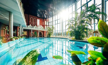 Helles, geräumiges Schwimmbad mit großen Fenstern und üppigen Pflanzen. Liegestühle stehen am Rand, während Sonnenlicht die Wasseroberfläche erhellt.