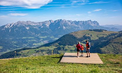 Drei Personen stehen auf einem Holzsteg und betrachten eine malerische Alpenlandschaft mit grünen Wiesen und majestätischen Bergen im Hintergrund.
