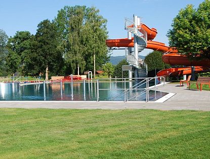 Ein Freibad mit einer großen Rutsche und einem Schwimmbecken, umgeben von grüner Wiese und Bäumen, bei sonnigem Wetter. Die Anlage wirkt einladend und gepflegt.