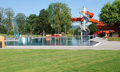Ein Freibad mit einer großen Rutsche und einem Schwimmbecken, umgeben von grüner Wiese und Bäumen, bei sonnigem Wetter. Die Anlage wirkt einladend und gepflegt.