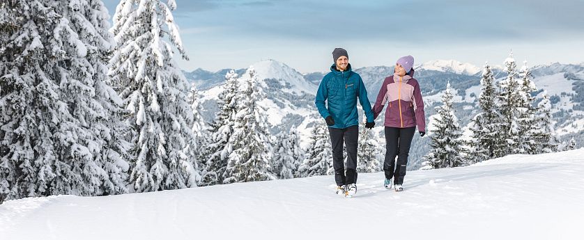 Ein Paar wandert im Winter durch schneebedeckte Berge. Sie tragen warme, bunte Kleidung und Mützen. Der Himmel ist klar, und die Landschaft zeigt verschneite Tannen.