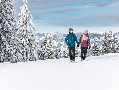 Ein Paar wandert im Winter durch schneebedeckte Berge. Sie tragen warme, bunte Kleidung und Mützen. Der Himmel ist klar, und die Landschaft zeigt verschneite Tannen.