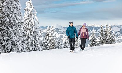 Ein Paar wandert im Winter durch schneebedeckte Berge. Sie tragen warme, bunte Kleidung und Mützen. Der Himmel ist klar, und die Landschaft zeigt verschneite Tannen.