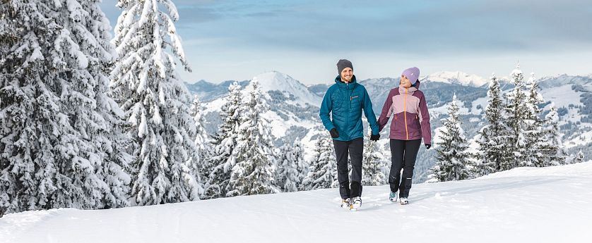 Ein Paar wandert im Winter über einen schneebedeckten Berg. Sie tragen warme Kleidung und halten sich an den Händen. Verschneite Bäume und Berge im Hintergrund.