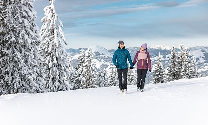 Ein Paar wandert im Winter über einen schneebedeckten Berg. Sie tragen warme Kleidung und halten sich an den Händen. Verschneite Bäume und Berge im Hintergrund.